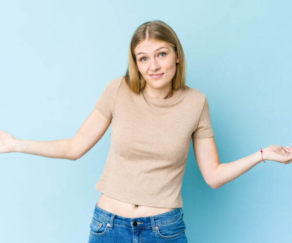 Young blonde woman isolated on blue background doubting and shrugging shoulders in questioning gesture.